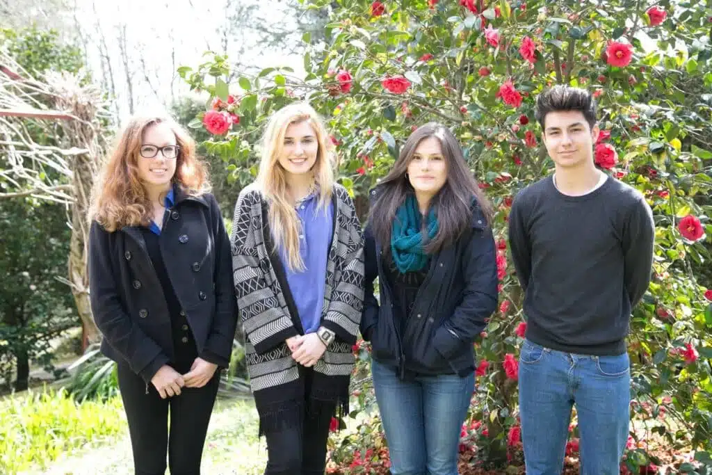 Cuatro jóvenes alumnos posando sonrientes en fila frente a un arbusto de camelias en flor en el jardín