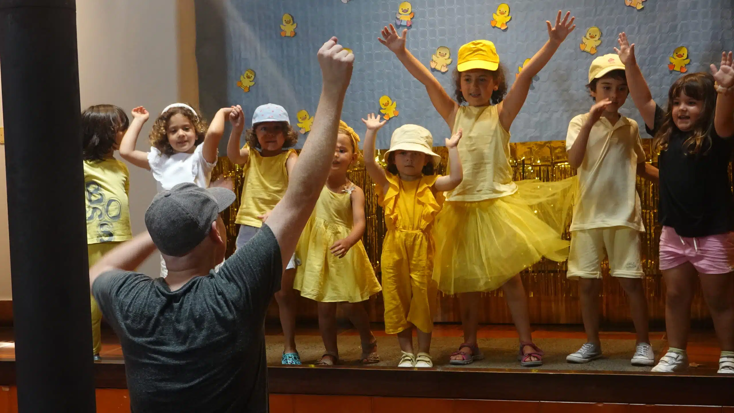 Alumnos pequeños disfrazados de amarillo bailando en el escenario dirigidos por un profesor