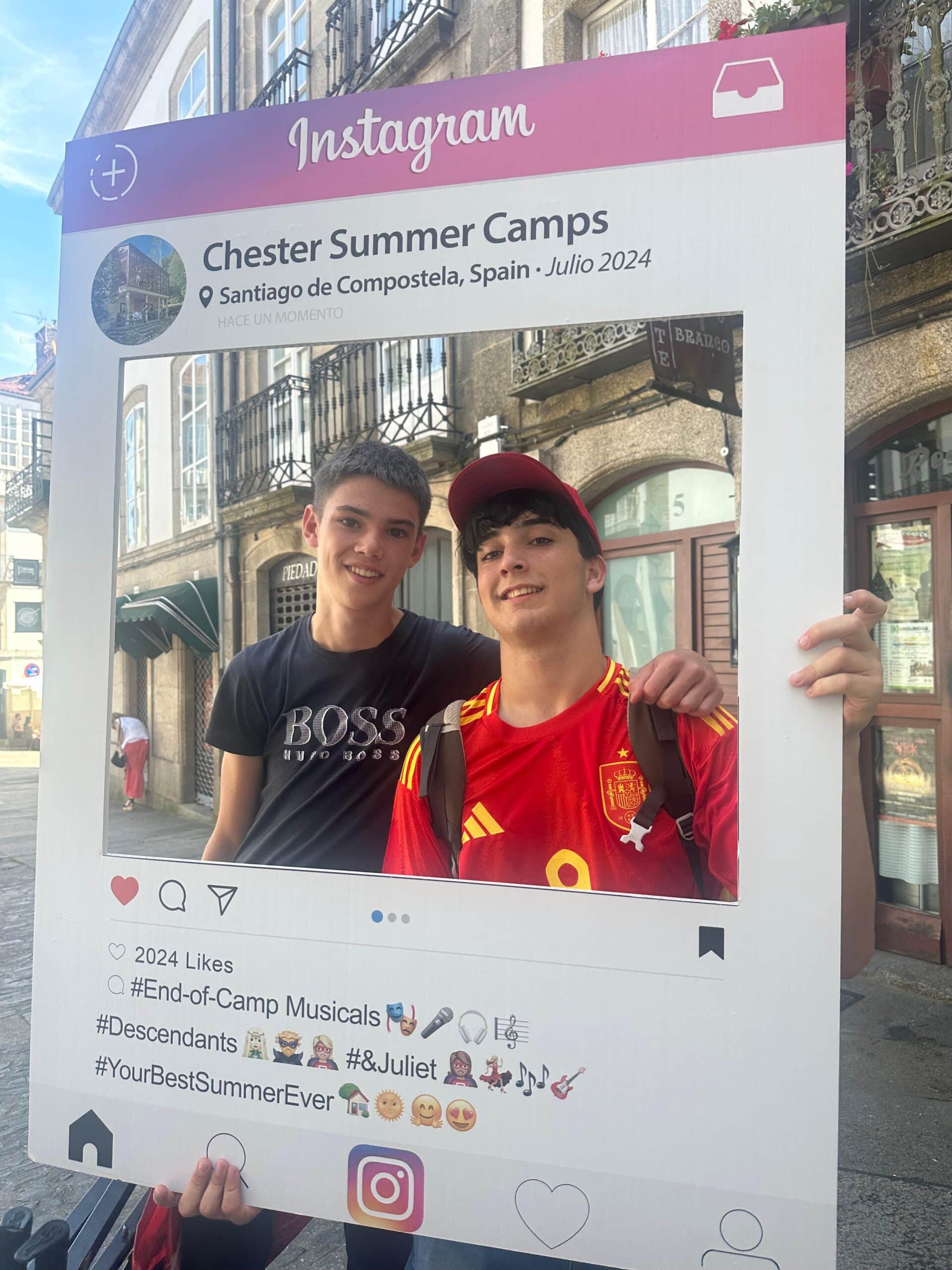 Dos jóvenes sonriendo detrás de un marco gigante que simula una publicación de Instagram de 'Chester Summer Camps' en Santiago de Compostela, julio de 2024
