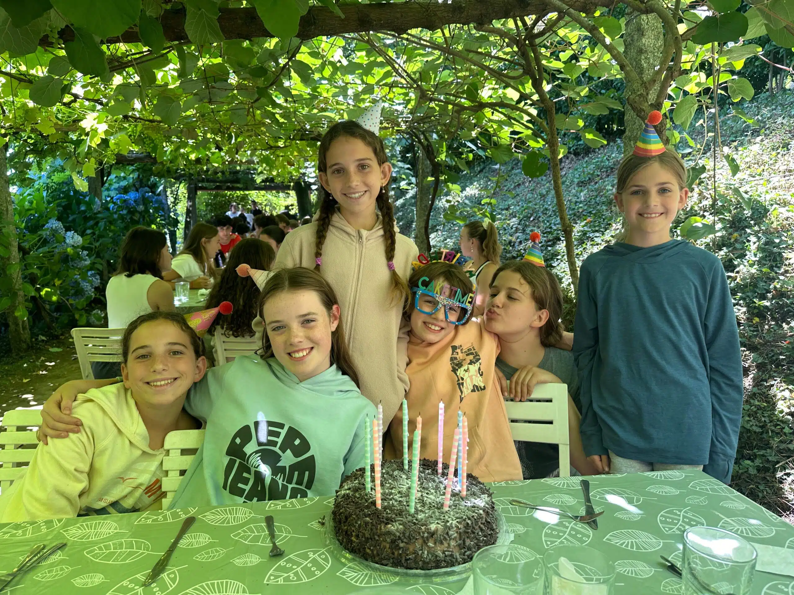 Grupo de alumnas sonriendo alrededor de una mesa con una tarta de cumpleaños y gorros de fiesta durante una celebración al aire libre