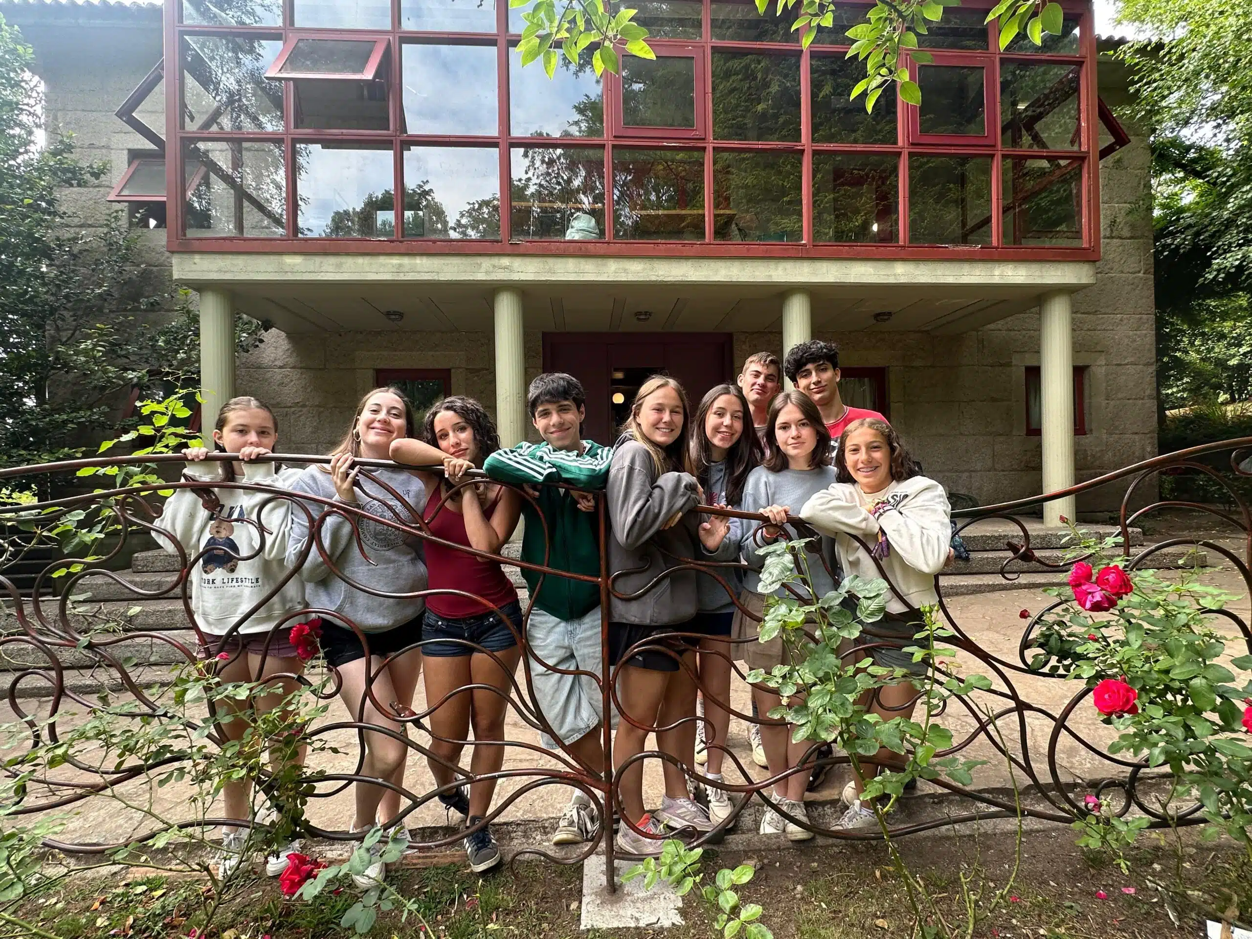 Grupo de alumnos adolescentes sonriendo y apoyados en una barandilla metálica en el jardín frente al edificio del colegio
