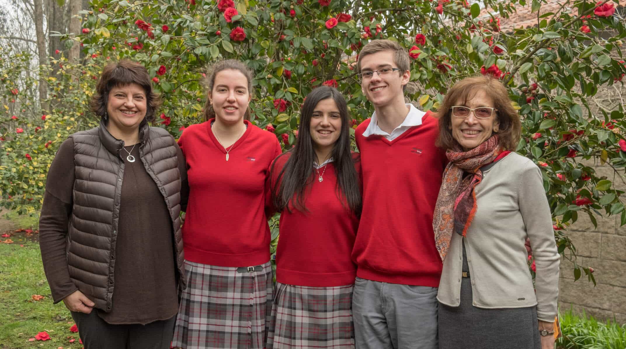 Dos profesoras y tres alumnos adolescentes con uniforme rojo sonriendo frente a un arbusto de camelias en flor