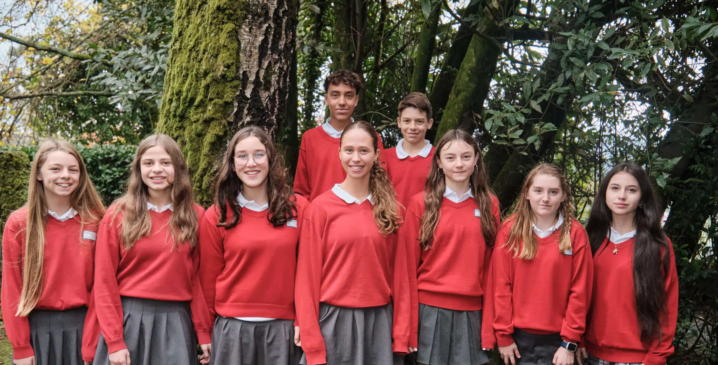 Grupo de nueve alumnos con uniforme escolar rojo posando sonrientes en dos filas frente a un gran árbol en el jardín
