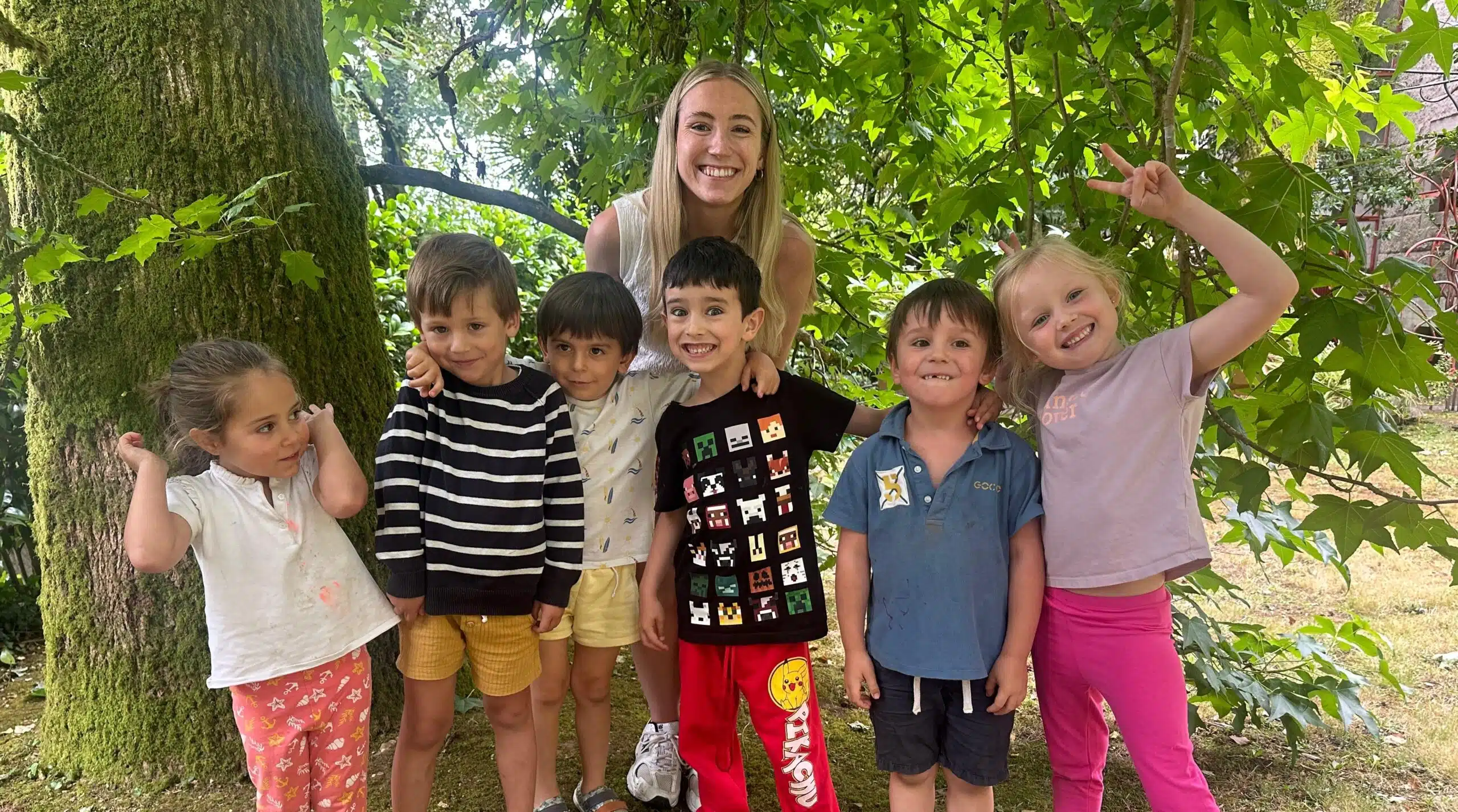 Profesora joven sonriendo y posando con un grupo de seis alumnos pequeños bajo un árbol en el jardín